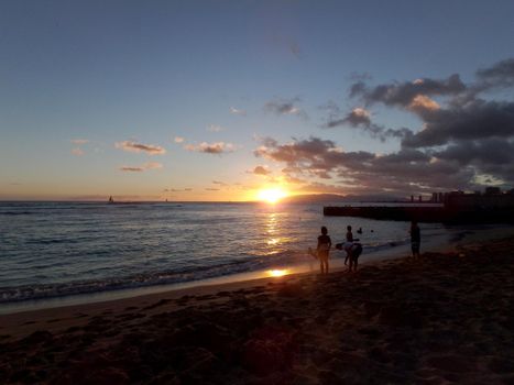 People watch dramatic Sunset on Kaimana Beach over the ocean with boats sailing on the water on Oahu, Hawaii.  July 2014.