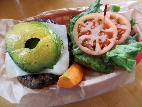 Open Swiss Cheese Hamburger with Veggies of Avocado, Lettuce, tomateos and Carrots in a red plastic basket on a table.