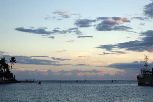 Opening to the sea at Kewalo Basin Harbor at dusk with people standing on shore watching sunset and boat docked at pier on Oahu, Hawaii.