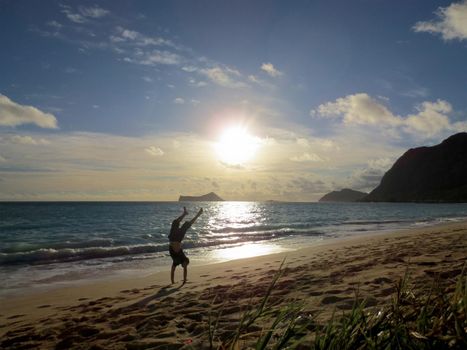 In Shape man does handstand wearing t-shirt and shorts on the Waimanalo beach on Oahu with his legs open during sunrise over the ocean with Rabbit Island, Rock Island, and Makapuu visible.