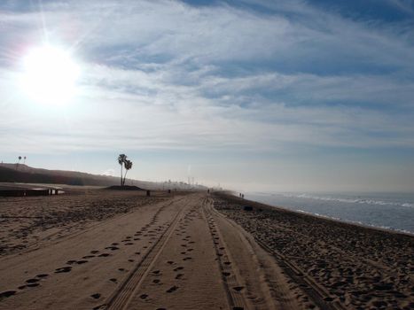 Tire tracks and footprints in the sand with palm trees and power plant in the distance with sun rising on Dockweiler Beach State Park, Playa Del Rey in LA, California.  January 2014.