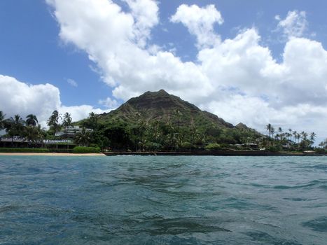 Beach and Makalei Beach Park with seawall, coconut trees, homes, and Diamond Head Crater in the distance and clouds in the sky on Oahu, Hawaii viewed from the water. 