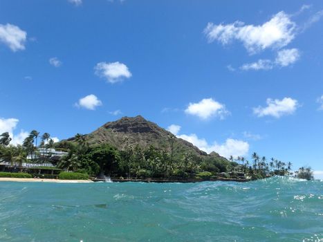 Beach and Makalei Beach Park with seawall, coconut trees, homes, and Diamond Head Crater in the distance on Oahu, Hawaii viewed from the water.