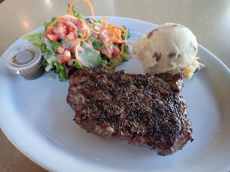 New York Steak, Mashed Potato, and Salad on white plate with dressing on a table.