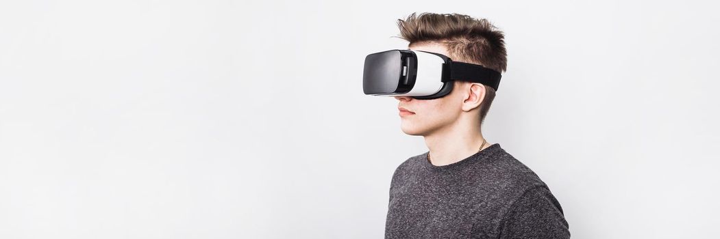 A young guy, a teenager stands in a villard vr glasses on a white background. Augmented reality in our time. A virtual reality glasses boy plays games and learns at school. Web banner