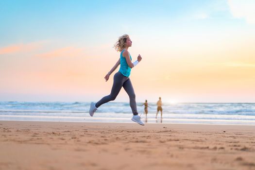 Active sporty woman run along ocean surf by water pool to keep fit and health. Sunset sand beach background with sun. Woman fitness, jogging workout and sport activity on summer family holiday.