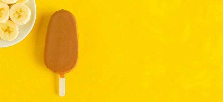 Banana vanilla popsicle in chocolated glaze on bright yellow background.Slices of banana on the plate,copyspace.Summer cool dessert.