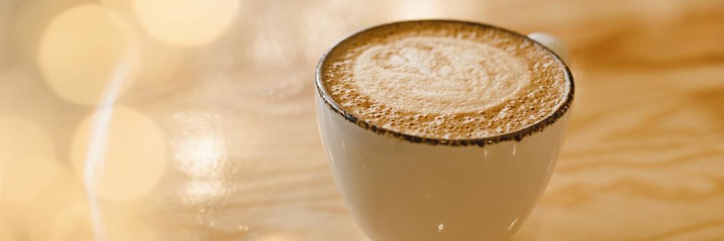 Mug of hot cappuccino on a wooden table in a cafe with yellow garland lights. Web banner.