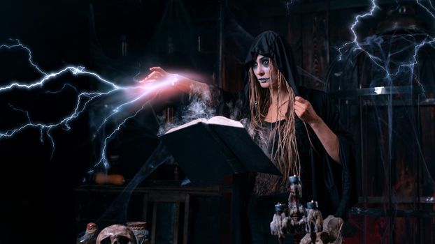 Halloween concept. Witch dressed black hood standing dark dungeon room use magic book for conjuring magic spell with lightning. Female necromancer wizard gothic interior with skull, cage, spider web