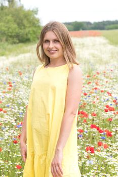 a beautiful young blonde woman in a yellow dress stands among a flowering field of poppies, daisies, cornflowers and laughs. High quality photo