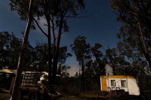 White caravan lit up by a full moon and makeshift homes from a small community in the woods. Trees in the Australian bush frame the night sky with clouds and stars. Mudgee, New South Wales