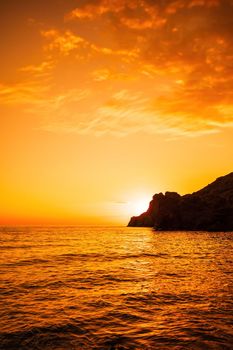 A red burning sunset with the silhouette of a cliff over the sea. Abstract nature summer or spring ocean sea background. Small waves on golden warm water surface with bokeh lights from sun.