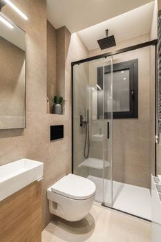 Simple bathroom with black shower, white toilet, wooden furniture and beige tiles. Modern interior of refurbushed apartment