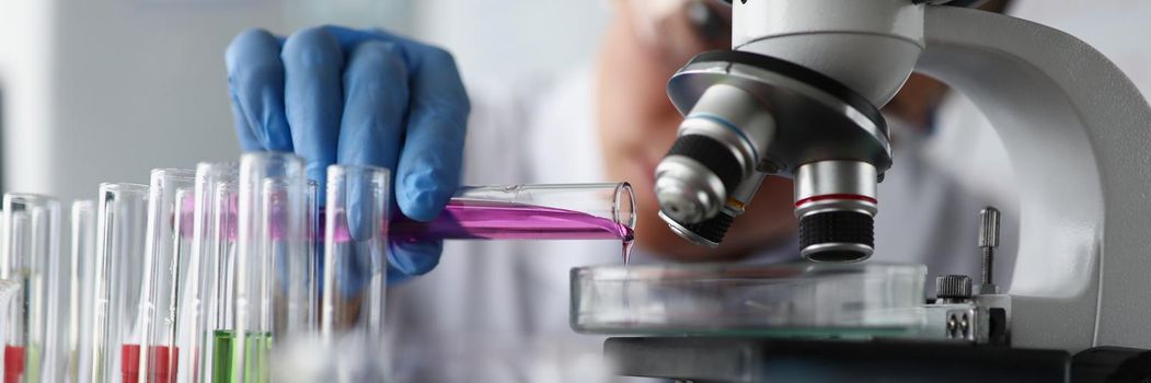 Close-up of female scientist pouring pink sample liquid on glass container under microscope in lab. Smart chemist or biologist in office. Science concept