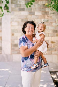 Grandmother stands holding a little girl in her arms near a stone wall. High quality photo