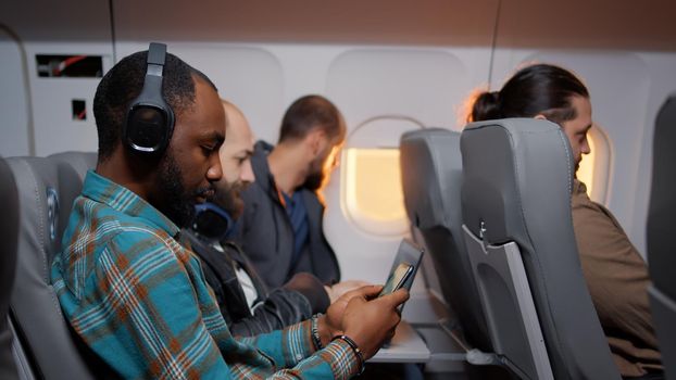 Male tourist using smartphone on airplane flight during sunset, browsing internet to pass time travelling to holiday destination. Flying in economy class and using phone.