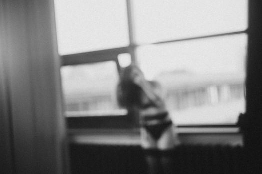 Gorgeous woman wearing black lingerie indoors. Portrait of a beautiful dreamy girl in a black underwear by the window. Black and white photo