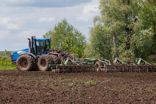 Blue New Holland tractor with double wheels pulling disc harrow with roller basket at hot sunny day in Tula, Russia - June 4, 2022