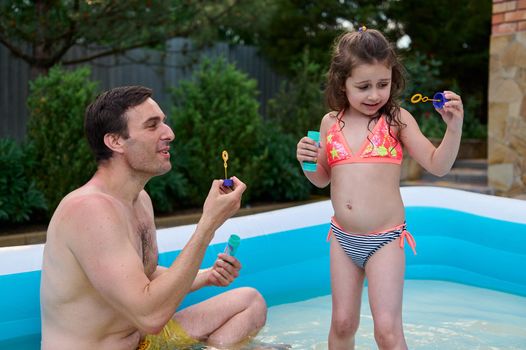 Adorable child, baby girl in bathing suit blows soaps bubbles in the inflatable swimming pool, next to her loving farher. Happy childhood. Family weekend together, love, togetherness. Father's Day