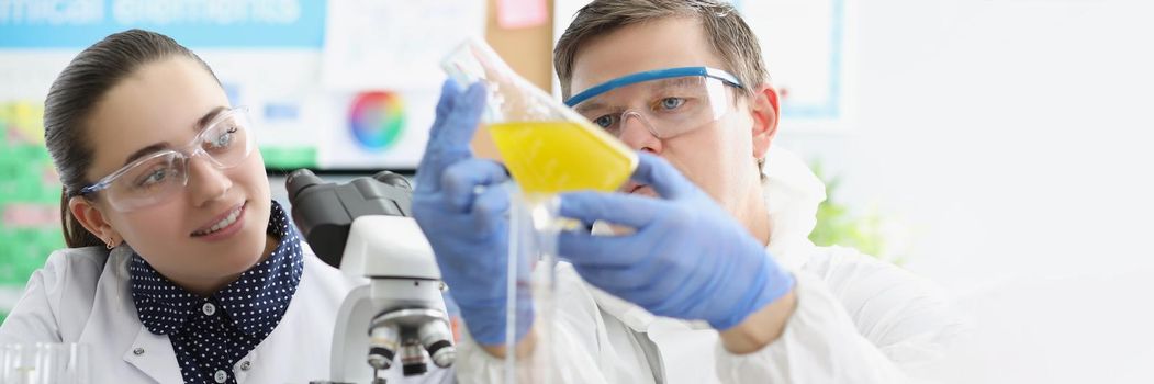 Portrait of laboratory workers man and woman examine yellow liquid in flask. Biologists use microscope for research. Chemistry, biology, science concept