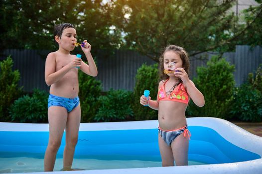 Caucasian funny kids, boy and girl, brother in sister, dressed in swimming trunks, blowing soap balls in the inflatable water pool on summer vacation. Water fun for children on a hot summer days