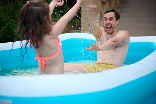 Loving father and his cute little daughter splashing each other with water, having fun together outdoors in the inflatable swimming pool on a warm sunny summer day. Family weekend, fatherhood