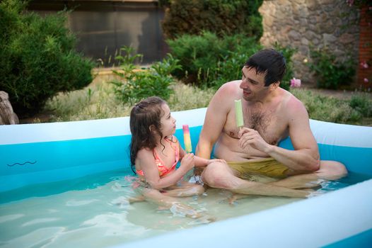 Cute little girl and her loving dad enjoying summer holidays together, sitting in an inflatable pool and eating sweet ice cream. Father and daughter spend weekend outdoors. Happy family relationships