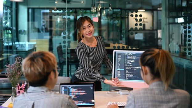 Confident female team leader and young software engineers working with programming code on computer screen in contemporary office.