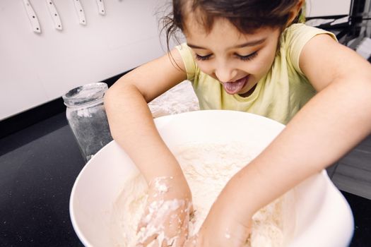 funny little girl with her tongue out mixing flour in a bowl to make homemade bread dough, healthy food concept at home