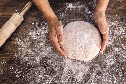hands of an unrecognizable person taking a rustic bread dough from a wooden table full of flour, concept of healthy food at home