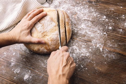 hands cutting a loaf of rustic bread in slices on a wooden table full of flour, concept of healthy food at home