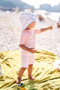 Little girl in a panama hat stands on the beach, pointing her finger into the distance. High quality photo