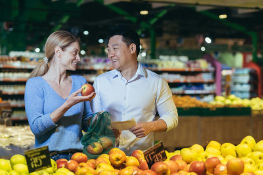happy mixed race asian couple family man and woman choosing fruits or vegetables in grocery store supermarket. Joint daily shopping together. Buyers customer select product pick an apple in eco bag