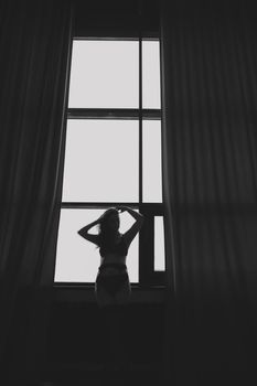 Gorgeous woman wearing black lingerie indoors. Portrait of a beautiful dreamy girl in a black underwear by the window. Black and white photo