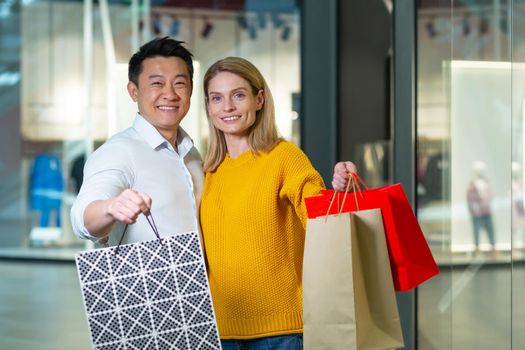 Young asian man and woman couple in supermarket shopping, looking at camera and smiling, holding colored packages with goods