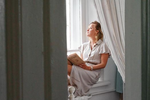 A middle-aged woman in a cream dress sits mysteriously and looks out the window on the windowsill. Green trees outside