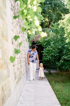 Grandmother with her little granddaughter walk holding hands against a stone wall in the garden. High quality photo