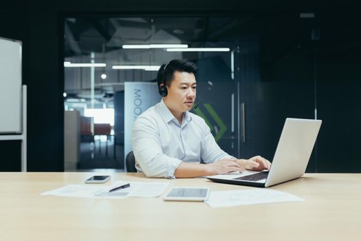 Young handsome Asian businessman working on laptop in headphones with microphone. Sitting at a desk in a modern office. Serious, focused.