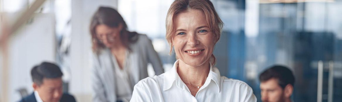 Portrait of beautiful smiling mature businesswoman with her colleagues on background in office.