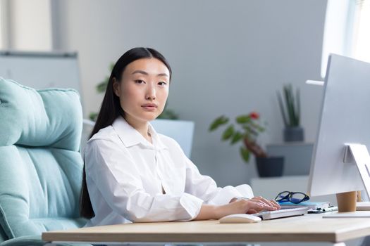 Close-up photo. Portrait of a young beautiful Asian woman secretary sitting at the desk at the computer in the office in business clothes, looking at the camera.