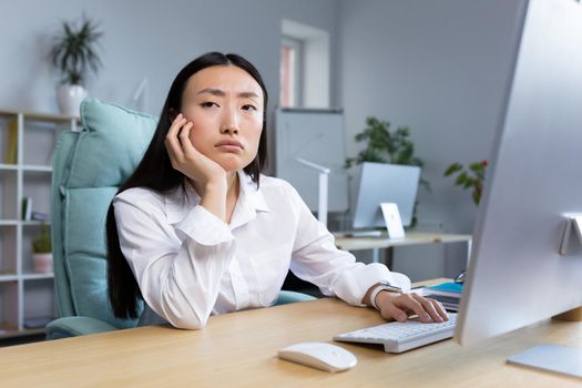 Boring and monotonous work. A young beautiful Asian business woman, an office worker sits at a desk at a computer, is sad, bored, performs monotonous work. He holds his head with one hand.