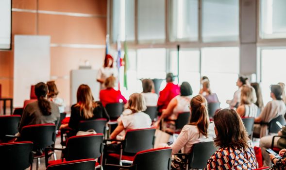 Business and entrepreneurship symposium. Female speaker giving a talk at business meeting. Audience in conference hall. Rear view of unrecognized participant in audience.