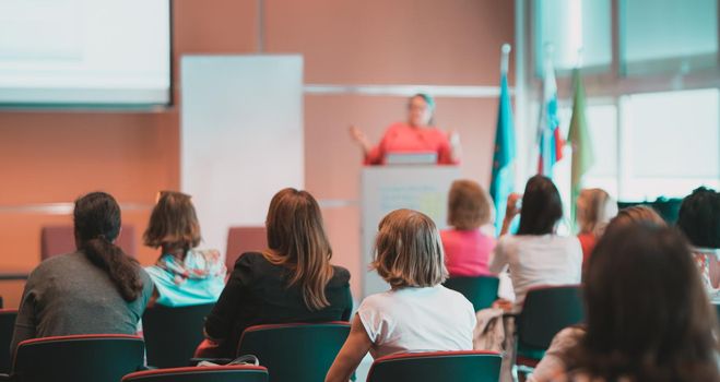 Business and entrepreneurship symposium. Female speaker giving a talk at business meeting. Audience in conference hall. Rear view of unrecognized participant in audience.