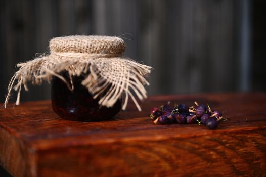 Close-up. Still life. A glass jar of homemade jam, with burlap on the lid tied with rope and ripe gooseberries scattered on a rustic wood surface. Canning, sweet preserves.