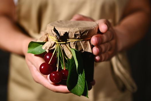 Details: Hands of a woman confectioner in a beige chef apron, handing a jar of homemade jam of ripe fresh cherries. Canning, preserved sweet food.