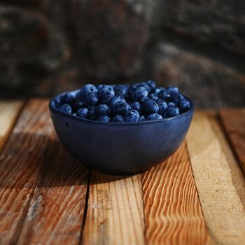 Still life composition of fresh sweet ready-to-eat blueberries from organic farm in a blue ceramic bowl standing on a wooden crate against gray stone background. Copy space for advertising text