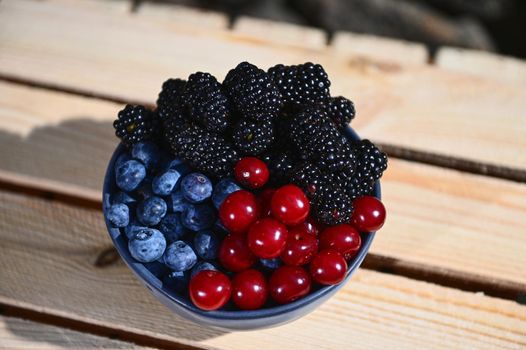 View from above of fresh juicy and ripe seasonal wild berries from an organic farm- blueberries, blackberries and cherries in a blue ceramic bowl on wooden crate. Web banner, copy ad space for text