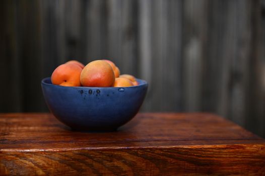 Ripe ready-to-eat corlorful apricot fruits in a blue ceramic bowl standing on a wooden surface against rustic wood background