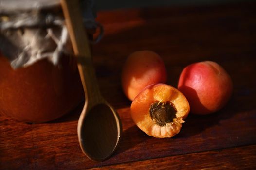Soft focus on halves of sweet, ripe, red, sunny, juicy and ready-to-eat apricot with pit next to a wooden tea spoon and fruit jam jar on a rustic wooden surface