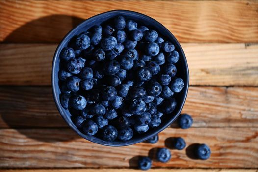 Flat lay. Horizontal composition of a blue ceramic bowl of fresh, ripe and ready-to-eat blueberries on a wooden crate background. Organic farm, healthy eating. Still life. Copy space for text. Banner